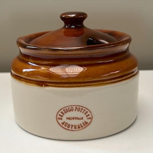 May include: A ceramic sugar bowl with a glossy brown lid and a cream-coloured base. The body features a circular stamp in brown ink that reads Bendigo Pottery Heritage Australia. The lid has a small knob handle and a notch for a spoon.