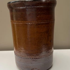 May include: A cylindrical stoneware crock featuring a glossy dark brown glaze on the upper portion and a lighter, muted greyish-brown tone on the base. Two horizontal bands of embossed circular patterns encircle the body, and a maker's mark is visible.
