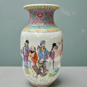 May include: A white ceramic vase with a flared rim and a wide body. The vase features a colorful floral design on the rim and a painted scene of figures in traditional clothing. The vase is approximately 30 cm tall.