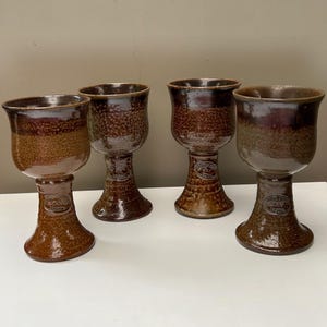 以下が含まれることがあります： 光沢のある仕上げと広がった縁を持つ4つの茶色の陶器製ゴブレット。各ゴブレットはテクスチャ加工されたベースと、ステムにBendigo Pottery Epsomと書かれた小さな楕円形のスタンプが特徴です。釉薬は濃い茶色から明るい茶色へと変化しています。
