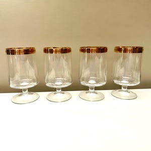 May include: Four clear glass goblets with a gold rim. The goblets have a short stem and a wide, cylindrical bowl. The gold rim adds a touch of elegance to the glassware. The goblets are arranged in a row on a white surface.