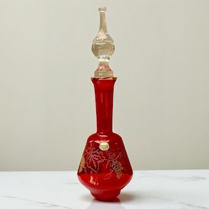 May include: A tall, red glass bottle with a clear stopper. The bottle has gold grape and leaf designs. The stopper is clear and bulbous. The bottle is likely for perfume or liquids. The bottle's height is approximately 25 cm.