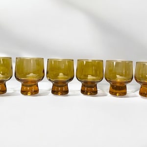 May include: Six amber-colored, stemless glasses with a rounded bowl and a short, sturdy base. The glasses are arranged in a row, showcasing their vintage design and warm hue, perfect for serving beverages.
