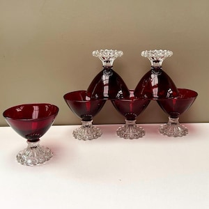 May include: A set of five ruby red glass dessert bowls with clear, textured, beaded pedestal bases. The bowls feature a wide, smooth, conical shape and are arranged with one bowl standing alone and four stacked in a pyramid formation.