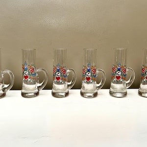 May include: Six clear glass shot glasses with handles. Each glass is decorated with a floral design in red, blue, and yellow, with a red heart at the centre. The glasses are arranged on a white surface.