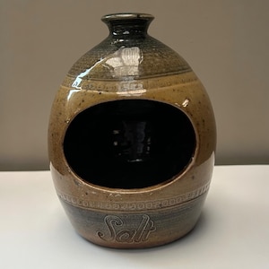 May include: A ceramic salt pig with a wide oval opening on the front. The vessel features a two-tone glaze with a tan speckled centre and dark brown bands at the top and bottom. The word Salt is embossed in a cursive script near the base.
