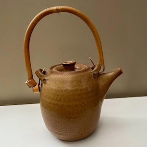 Peut inclure: Une théière en céramique beige avec une finition mouchetée, dotée d'un corps arrondi, d'un bec court et d'un couvercle avec un petit bouton. La théière est équipée d'une anse haute et arquée en bambou clair, fixée à deux boucles latérales.