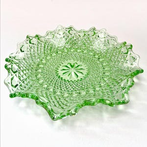 May include: A light green pressed glass plate featuring a detailed diamond pattern across the surface, a central starburst motif, and a decorative scalloped edge. The translucent material catches the light, highlighting the intricate geometric texture.