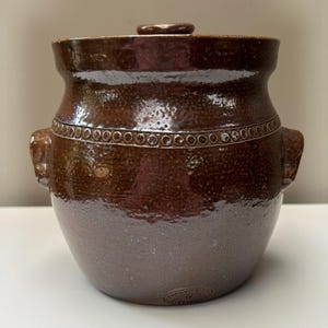 May include: A brown glazed ceramic crock with a lid, featuring two side handles and a decorative band of small circular indentations around the upper shoulder. The surface has a mottled texture, and a small circular maker's mark is visible at the base.