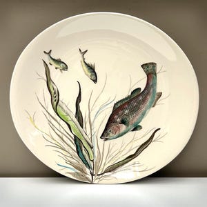 May include: A round, off-white ceramic plate featuring a detailed illustration of a large fish swimming among green seaweed, accompanied by two smaller fish in the upper left area. The artwork uses muted tones of green, brown, and blue.