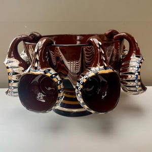 May include: A dark brown ceramic punch bowl with six attached mugs. The mugs have curved handles and are decorated with blue, white, and yellow patterns. The bowl has a decorative rim and a striped base.