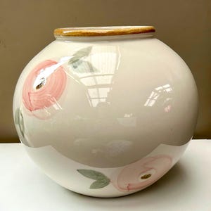 May include: A round, cream-coloured ceramic vase with a glossy finish. The vase features hand-painted, soft pink floral motifs with muted green leaves scattered across the surface. It has a distinct, thick, tan-coloured rim around the opening.