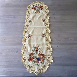 May include: Cream-colored embroidered table runner with a scalloped lace edge. The runner features floral and bunny designs in the center. The bunnies are depicted in a playful scene with baskets and flowers.