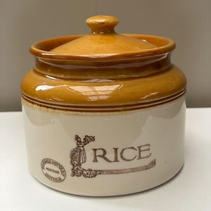 May include: A cylindrical ceramic storage jar with a cream-colored base and a glossy honey-brown lid. The front features brown text reading RICE, a decorative illustration of a person, and a circular stamp labeled Bendigo Pottery Heritage Australia.