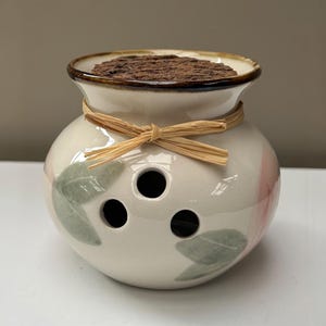 以下が含まれることがあります： 茶色の縁取りがあるクリーム色の丸い陶器製の壺で、前面に3つの円形の開口部と緑色の葉の模様が描かれています。首元には薄茶色のラフィアのリボンが結ばれており、上部には濃い茶色の質感のある円形の蓋が乗っています。