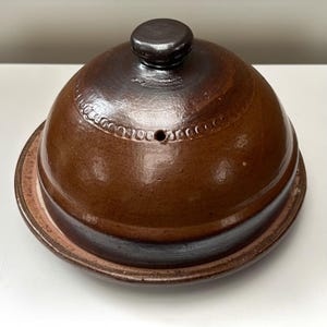 May include: A round, brown glazed ceramic cheese dome with a matching base plate. The dome features a dark brown knob handle at the top, a small circular steam vent, and a decorative embossed band of small circles encircling the upper section.