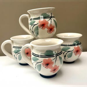 May include: A set of four cream-coloured ceramic mugs featuring a hand-painted floral pattern with soft pink blossoms and muted green leaves. Each mug has a rounded body, a curved handle, and two thin dark green horizontal bands encircling the rim.
