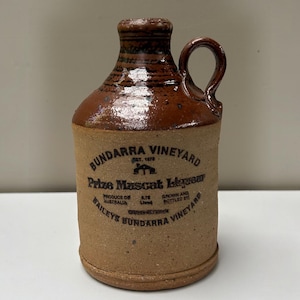 May include: A brown ceramic jug with a handle and a glazed neck. The body features black text reading BUNDARRA VINEYARD, Prize Muscat Liqueur, PRODUCE OF AUSTRALIA, 0.75 Litres, GROWN AND BOTTLED BY BAILEYS BUNDARRA VINEYARD, and an icon of a small house.