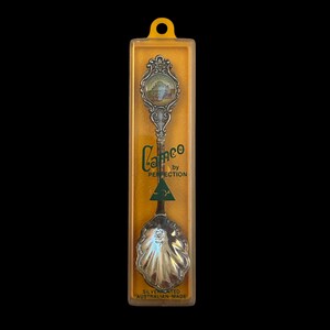 May include: A silver-plated souvenir spoon in a clear plastic case with a yellow backing. The spoon has a decorative handle with a small image and a shell-shaped bowl. The packaging reads "Cameo by Perfection" and "Australian Made."