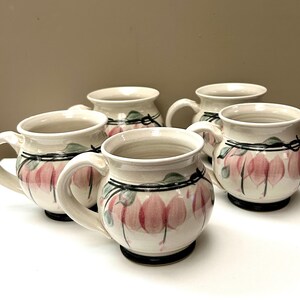 May include: A set of four round ceramic mugs with a cream-coloured glaze, each featuring a hand-painted pattern of pink bleeding heart flowers with green leaves and black stems. The mugs have a wide, curved body, a sturdy handle, and a dark base.