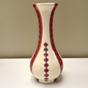 May include: A tall, cream-coloured ceramic vase with a flared rim and a rounded base. The surface is decorated with vertical bands of intricate red and black geometric patterns that run along the sides and down the centre of the body.