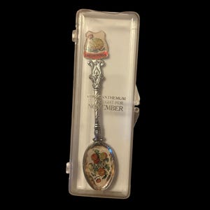 May include: A decorative silver spoon with a floral design and the text "CHRYSANTHEMUM" and "NOVEMBER". The spoon is in a clear plastic case. The spoon's bowl features a colourful floral design.