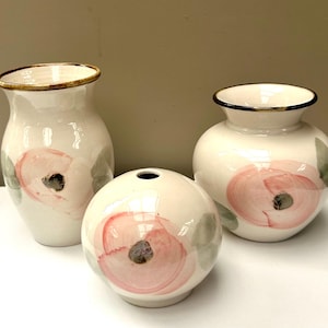May include: Three ceramic vases with a cream-coloured glaze, each featuring hand-painted pink floral motifs with dark centres and soft green leaves. The collection includes one tall slender vase, one rounded spherical vase, and one wider bulbous vase.