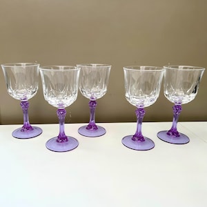 May include: Five clear crystal wine glasses with purple stems and bases. The glasses have a cut glass design on the bowl. The stems are ornate and the bases are round.