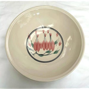 May include: A round, cream-coloured ceramic bowl featuring a central circular design. Inside the circle, there is a hand-painted illustration of pink, pod-like botanical elements with green leaves, all enclosed by a thin, dark blue circular border.