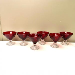 以下が含まれることがあります： ルビーレッドのシャンパンクーペグラス6個セット。各グラスは丸みを帯びたボウル型で、透明で質感のあるベースの上にあります。グラスは一列に並べられ、エレガントなデザインを際立たせています。