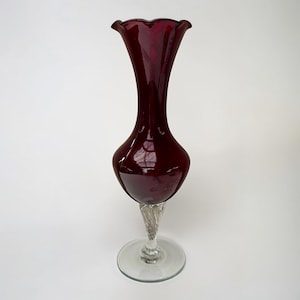 May include: A striking burgundy glass vase with a ruffled rim. The vase features a rounded body and a long, slender neck. It is supported by a clear, twisted glass stem and a circular base, presenting a classic design, perfect for floral arrangements.