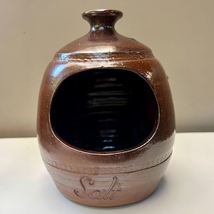 May include: A brown glazed ceramic salt pig with a wide front opening and a narrow neck at the top. The word Salt is embossed in cursive script on the lower front section. The surface has a smooth, glossy finish with subtle horizontal ridges.