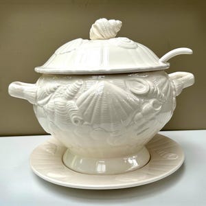 May include: A cream-coloured ceramic soup tureen with a matching lid and underplate. The tureen features a raised, embossed seashell pattern across its surface, two side handles, and a lid topped with a sculpted seashell finial.