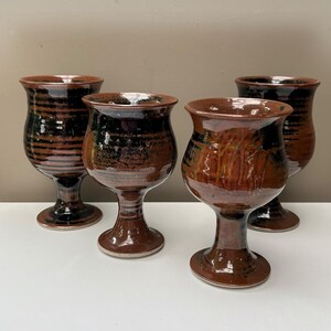 May include: A set of four ceramic goblets featuring a glossy glaze in shades of deep brown and black. Each goblet has a wide, flared rim, a rounded bowl, and a sturdy pedestal base. The surface displays horizontal ridges and mottled colour patterns.