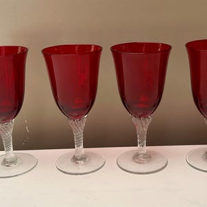 May include: Four ruby red wine glasses. Each glass has a clear, twisted stem and a round base. The bowls of the glasses are a deep red colour. The glasses are arranged in a row on a white surface.