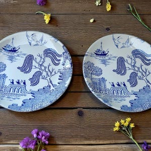 May include: Two white ceramic plates with a blue Willow pattern design. Each plate features a boat, birds, trees, and a bridge scene. The plates are set on a wooden surface with flowers.