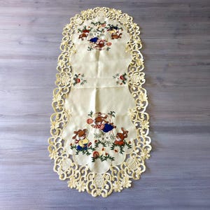May include: Cream-coloured, oval table runner with a scalloped, openwork lace border. The centre features embroidered cartoon bears in various outfits, playing in a garden setting. The runner is approximately 76 cm long.