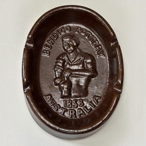 May include: An oval-shaped dark brown ceramic ashtray featuring a relief design of a person working with pottery. The text BENDIGO POTTERY is embossed at the top, and the text 1858 AUSTRALIA is embossed at the bottom of the central basin.