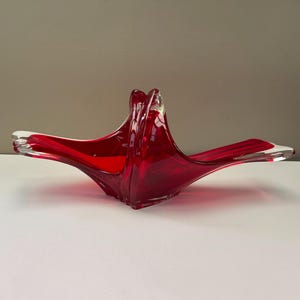 May include: A vibrant red glass basket featuring a clear glass handle and base. The vessel has an elongated, sweeping shape with pointed ends that curve upward, showcasing a smooth, translucent finish and a deep crimson colour throughout the body.