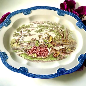 May include: Oval serving platter with a white centre and a blue scalloped border. The platter features a pastoral scene with a couple, trees, and a building. The border has decorative blue accents.