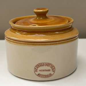 May include: A cylindrical ceramic sugar bowl with a glossy honey-brown lid and a cream-coloured base. The front features a circular embossed stamp that reads BENDIGO POTTERY HERITAGE AUSTRALIA. The lid has a small knob handle and a notch for a spoon.