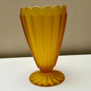 May include: A vintage amber-colored glass vase with a scalloped rim and a flared, ribbed body. The vase sits on a round, ridged base. The glass has a warm, translucent quality, and the design is reminiscent of Art Deco styles.