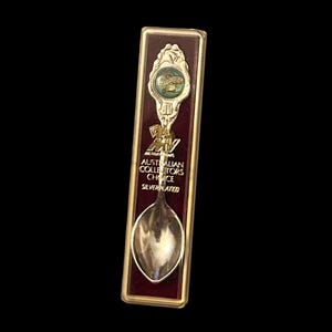 May include: A silver-plated souvenir spoon with a maroon background. The spoon has a decorative handle with a green and gold emblem and the text "Australian Collectors Choice". The spoon is encased in a rectangular gold-coloured frame.