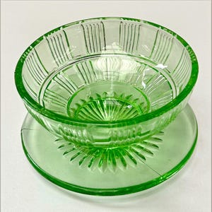 May include: A translucent light green glass bowl featuring a pressed pattern of vertical ribs along the upper rim and a sunburst design radiating from the centre of the base. The bowl sits atop a matching circular glass plate with a sunburst pattern.