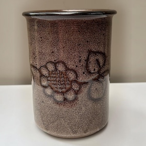 以下が含まれることがあります： 茶色とベージュの斑点模様の釉薬が施された円筒形の陶器製マグカップ。外側の表面には、中央の花と葉からなるエンボス加工のフローラルパターンが施されており、本体に対してより濃い茶色のトーンで表現されています。