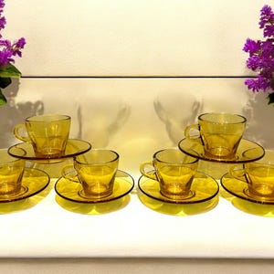 May include: A collection of eight amber glass teacups and saucers. The cups have a traditional design with handles, and the saucers are circular. The set is displayed on a white surface, with purple flowers in the background.