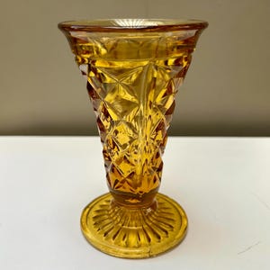 May include: Amber-colored glass vase with a diamond pattern and a round base. The vase has a flared top and a textured surface. The base has a ribbed design. The vase is made of glass and is likely vintage.