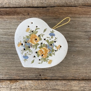 May include: A heart-shaped, white ceramic tea infuser with a floral design featuring yellow and blue flowers and green leaves. It has small holes and a gold cord for hanging. The infuser rests on a wooden surface.