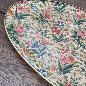 May include: Oval serving tray with a floral design. The tray has a cream-coloured background decorated with various flowers in shades of pink, red, yellow, and blue, with green leaves. It has a gold-coloured metal rim.