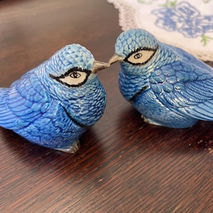 May include: Two ceramic blue bird ornaments with detailed feather textures and black and white eye accents. The birds are positioned beak-to-beak. The ornaments are on a dark wooden surface. The birds are a charming decorative item.